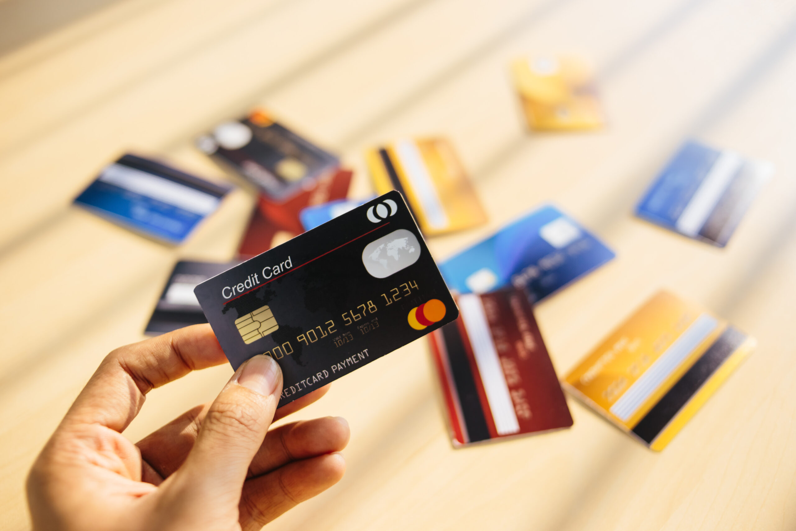 person holding a credit card in the foreground, with a pile of credit cards laying on a table in the blurred background